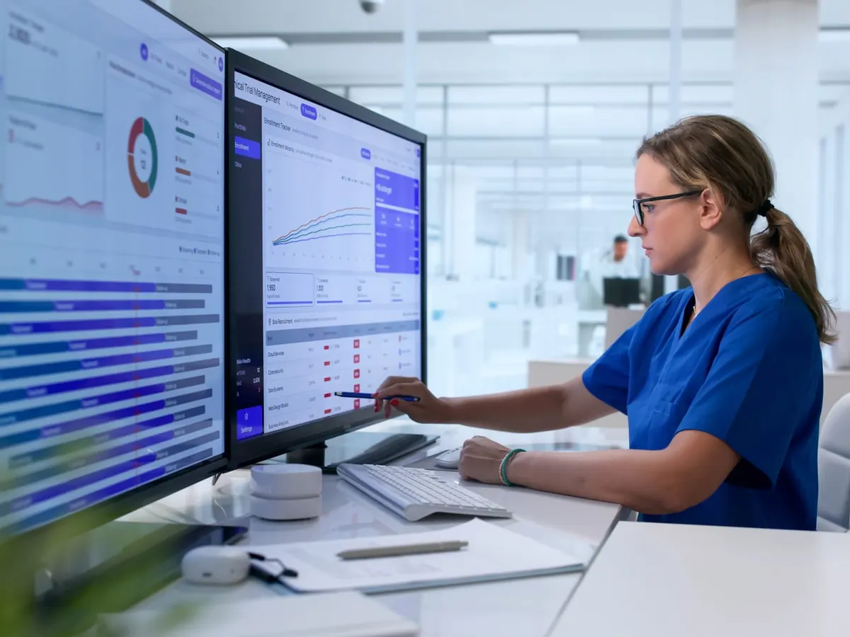 A woman in scrubs points to charts of data on two large monitors