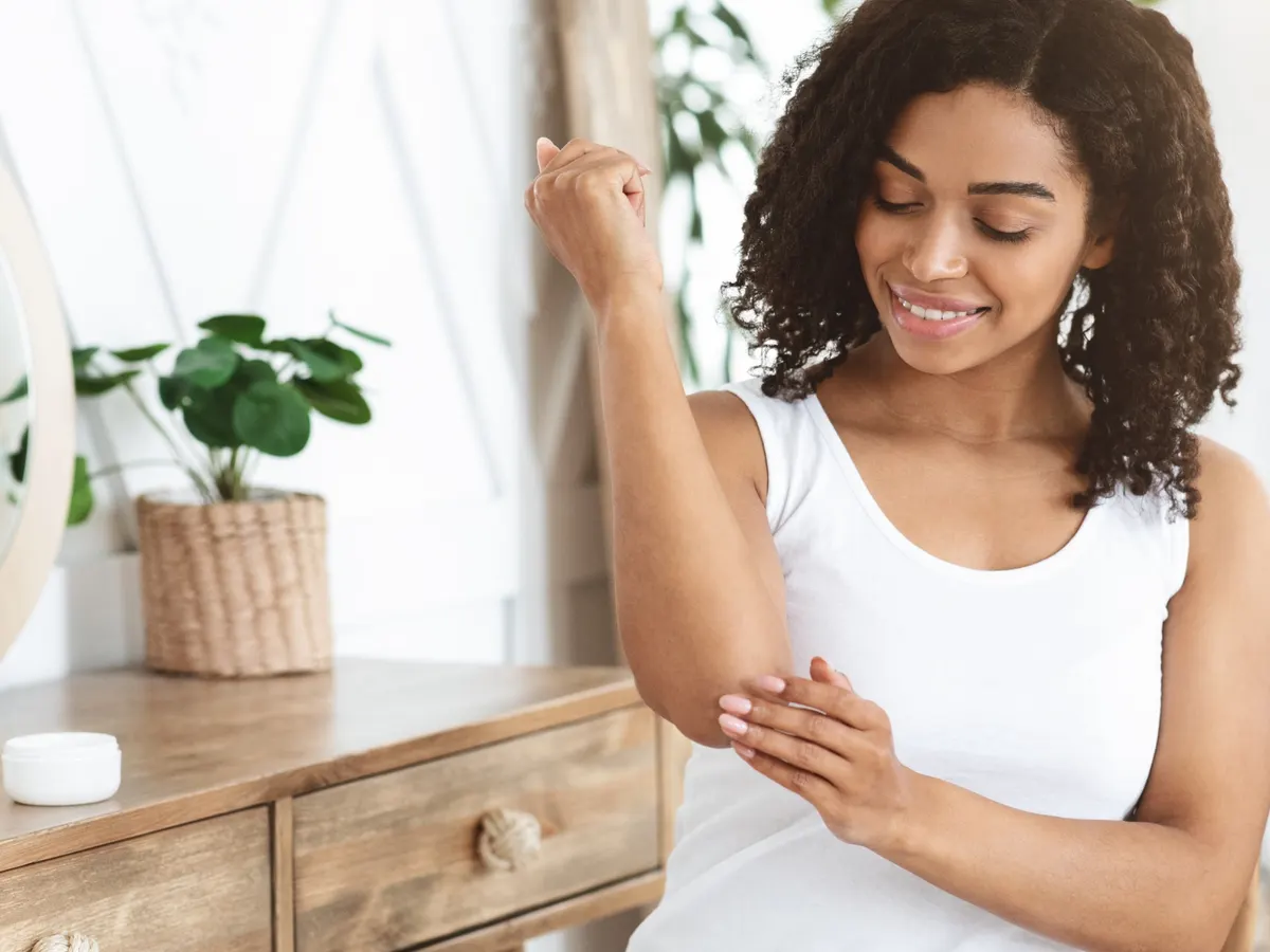 A smiling woman rubs cream into her elbow at a dressing table