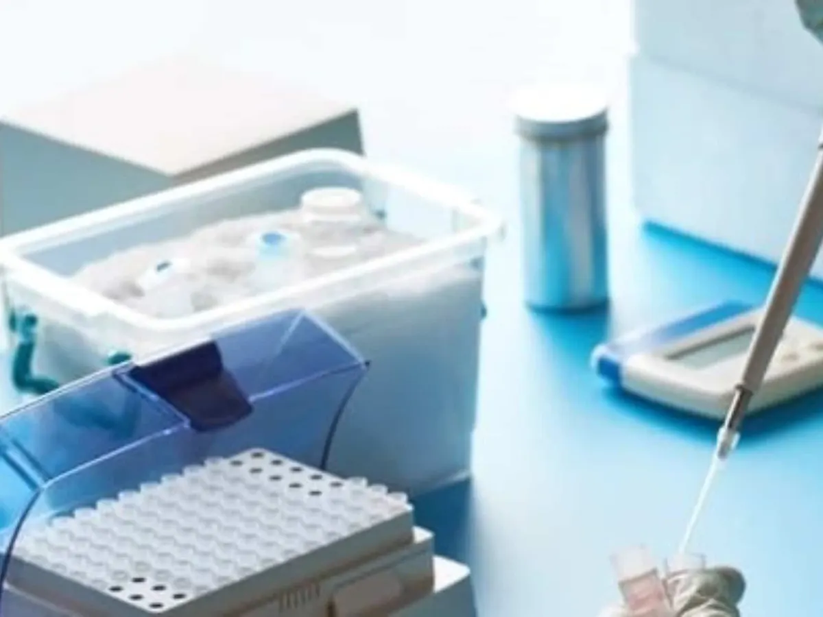 A gloved hand pipettes into a sample tube beside a box of vials and a rack of tubes