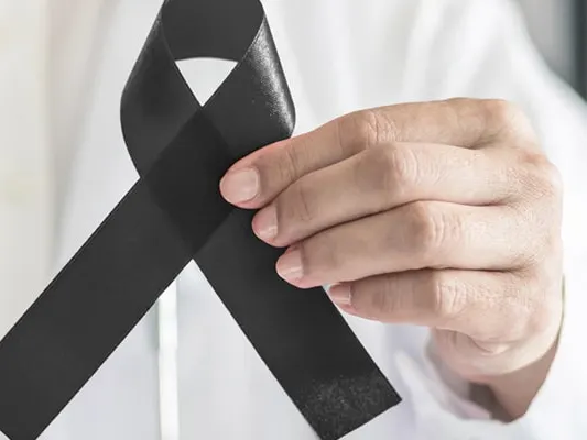 A hand holds up a black awareness ribbon in front of a white coat and stethoscope