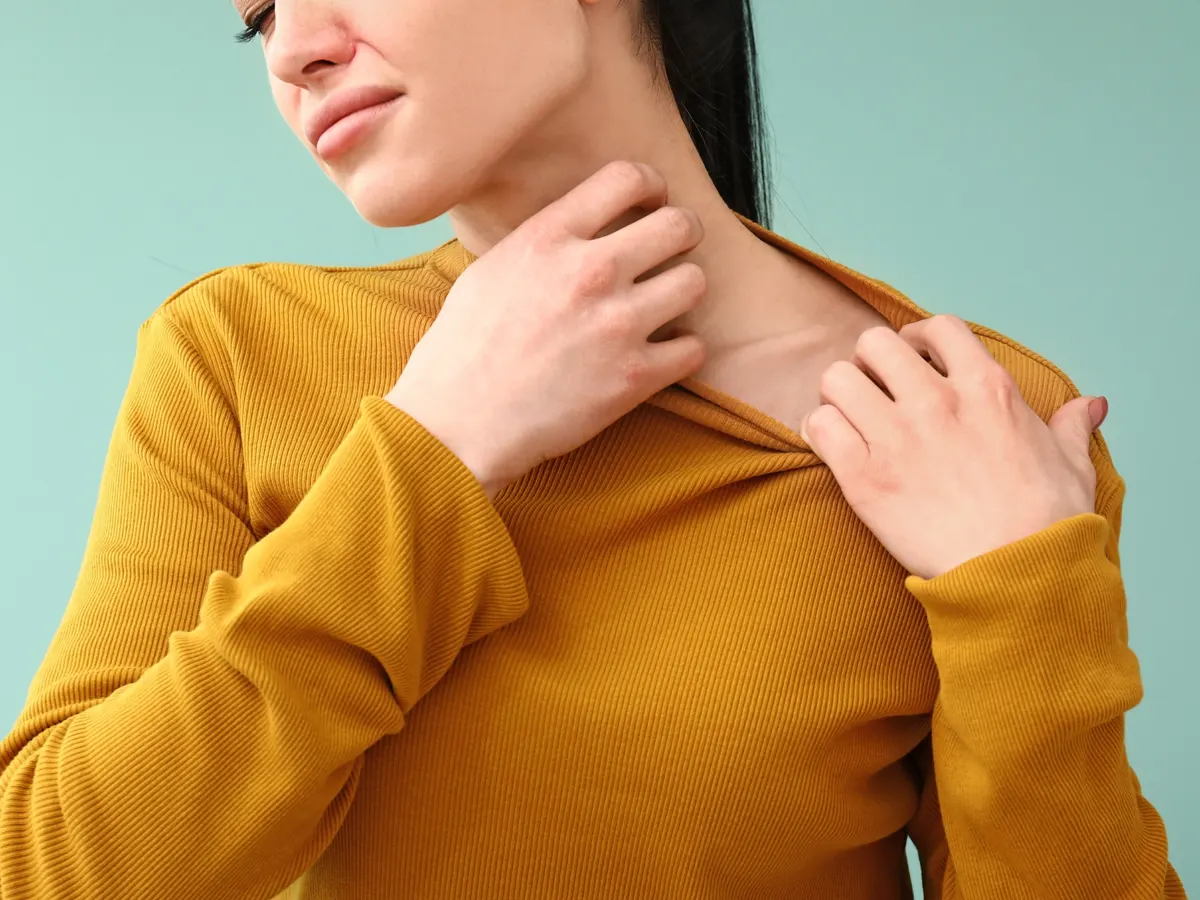 A woman in a mustard sweater scratches her neck and chest